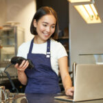 Smiling asian barista, girl with card terminal, payment machine and laptop, standing in cafe, processing payment for coffee order.