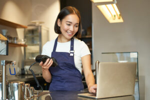 Smiling asian barista, girl with card terminal, payment machine and laptop, standing in cafe, processing payment for coffee order.